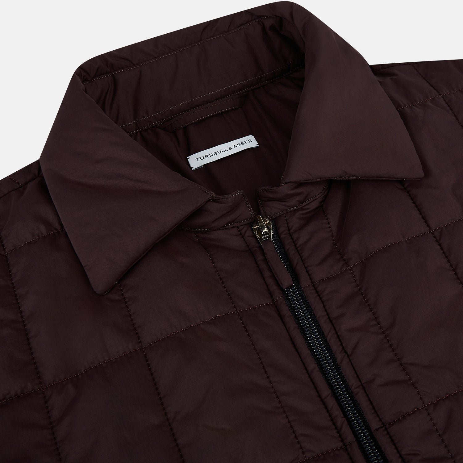 View 2 of product The Battersea Burgundy Quilted Jacket by Turnbull & Asser is a men's elegant outerwear featuring a front zipper, zippered side pockets, chest pocket, pointed collar, and buttoned cuffs, shown on a plain white background.