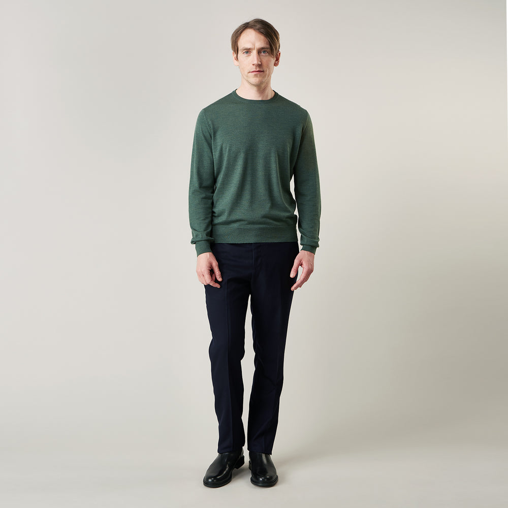 A man stands facing the camera against a plain light background, wearing the Turnbull & Asser Bottle Green Merino Kingston Crew Neck Jumper with dark pants and black shoes. His arms rest at his sides and he has a neutral expression.