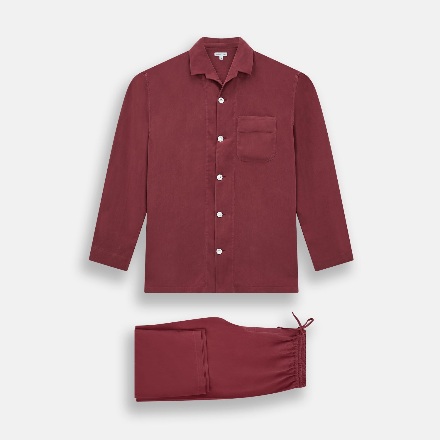 View 1 of product The Turnbull & Asser Burgundy Silk Pyjama Set features a long-sleeve, button-up shirt with chest pocket and matching pants with elastic waistband and side pocket, shown on a white background.