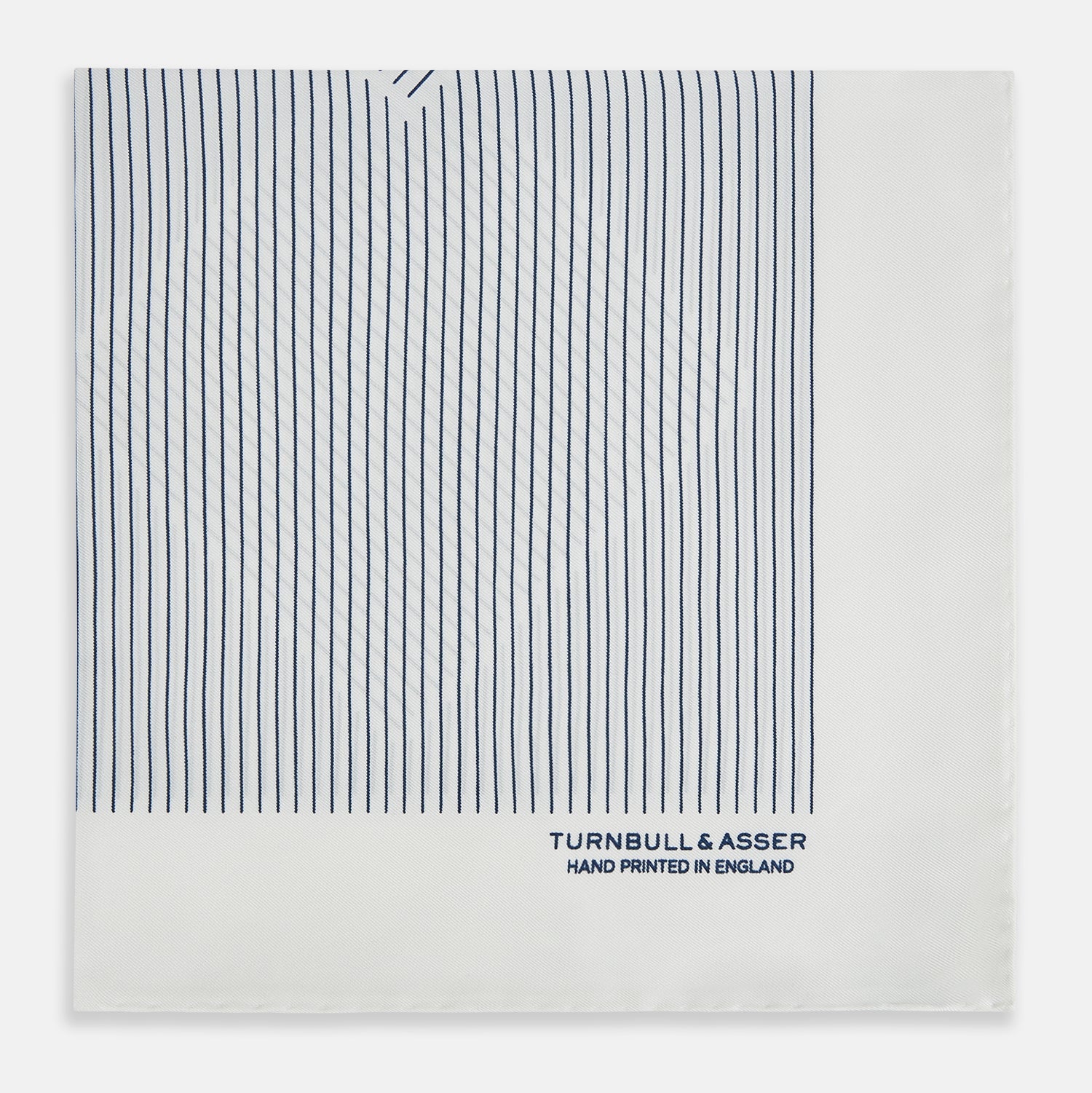 View 3 of product The White Stripe Silk Pocket Square by Turnbull & Asser features vertical navy pinstripes and “TURNBULL & ASSER HAND PRINTED IN ENGLAND” in blue at the lower right, with one corner neatly folded for a classic British look.