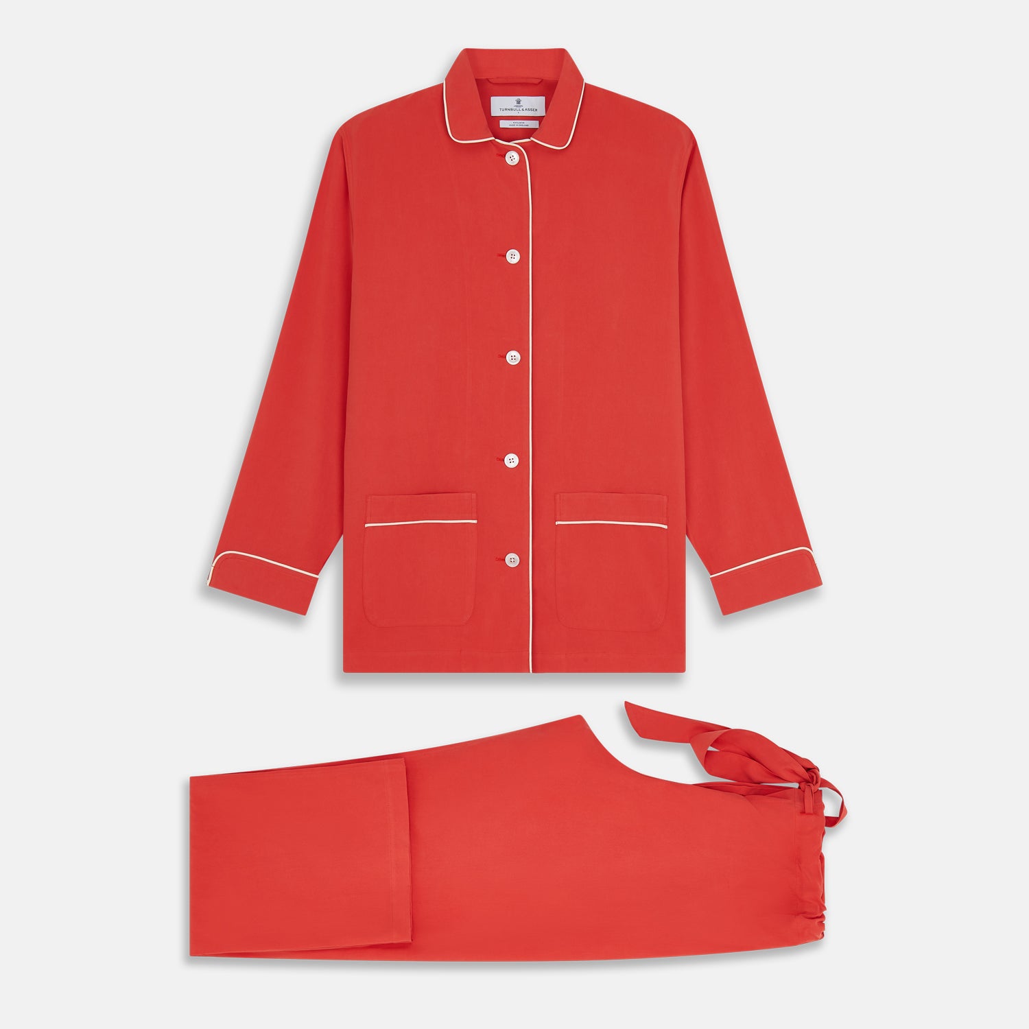 View 5 of product The Turnbull & Asser Women's Coral Pink Silk Thelma Pyjama Set features a long-sleeve button-up top with two front pockets and matching drawstring pants, shown on a white background.