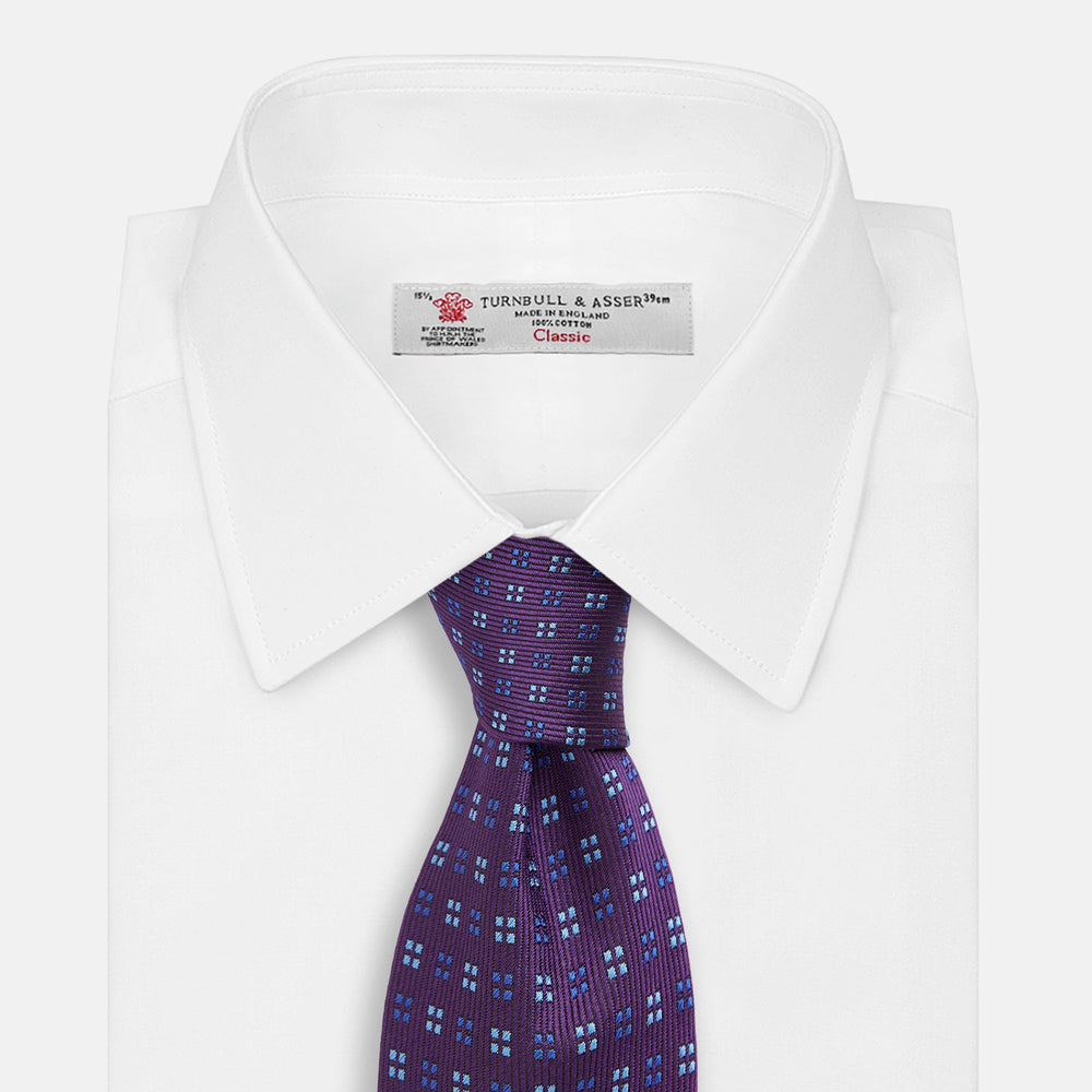 The Turnbull & Asser MINI SQUARE SPOT PLUM BLUE TIE is a luxury English silk necktie in plum, featuring a blue and white grid pattern of small squares, displayed rolled up against a simple light background.