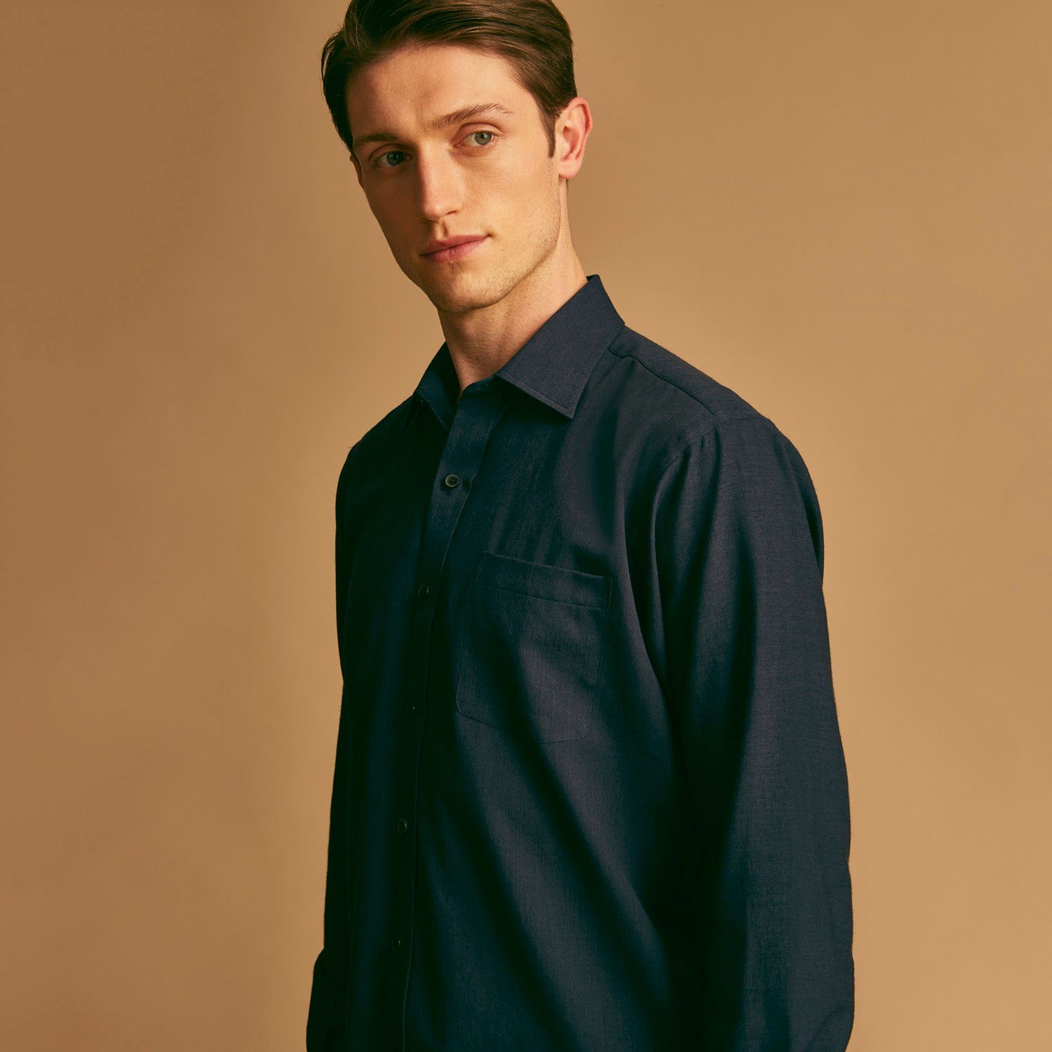 View 2 of product A man stands against a beige background wearing the Turnbull & Asser Navy Weekend Fit Cotton & Wool Shirt with a Derby collar and 1 button cuffs, paired with brown pants and black shoes, looking to the side with a neutral expression.
