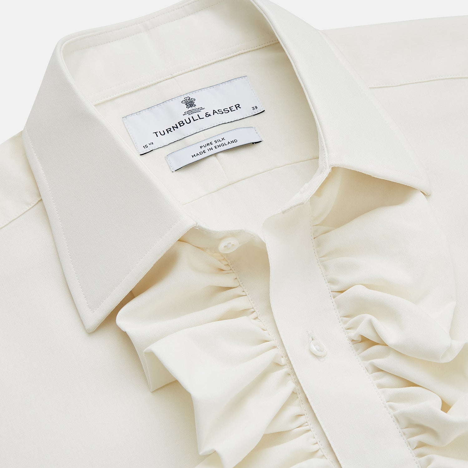 View 3 of product A man in a Cream Silk Regular Fit Ruffle-Front Wilfred Shirt by Turnbull & Asser stands against a dark background, one hand on a chair, gazing thoughtfully to the side.