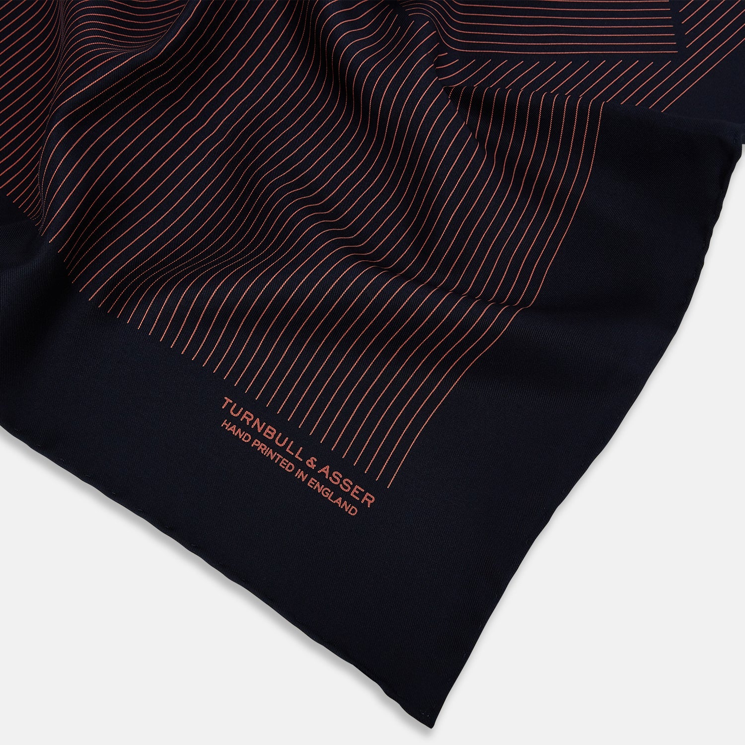 View 2 of product The Navy and Orange Stripe Silk Pocket Square by Turnbull & Asser is handmade in England from silk, featuring thin orange geometric stripes on navy and "Turnbull & Asser Hand Printed in England" printed in the corner.