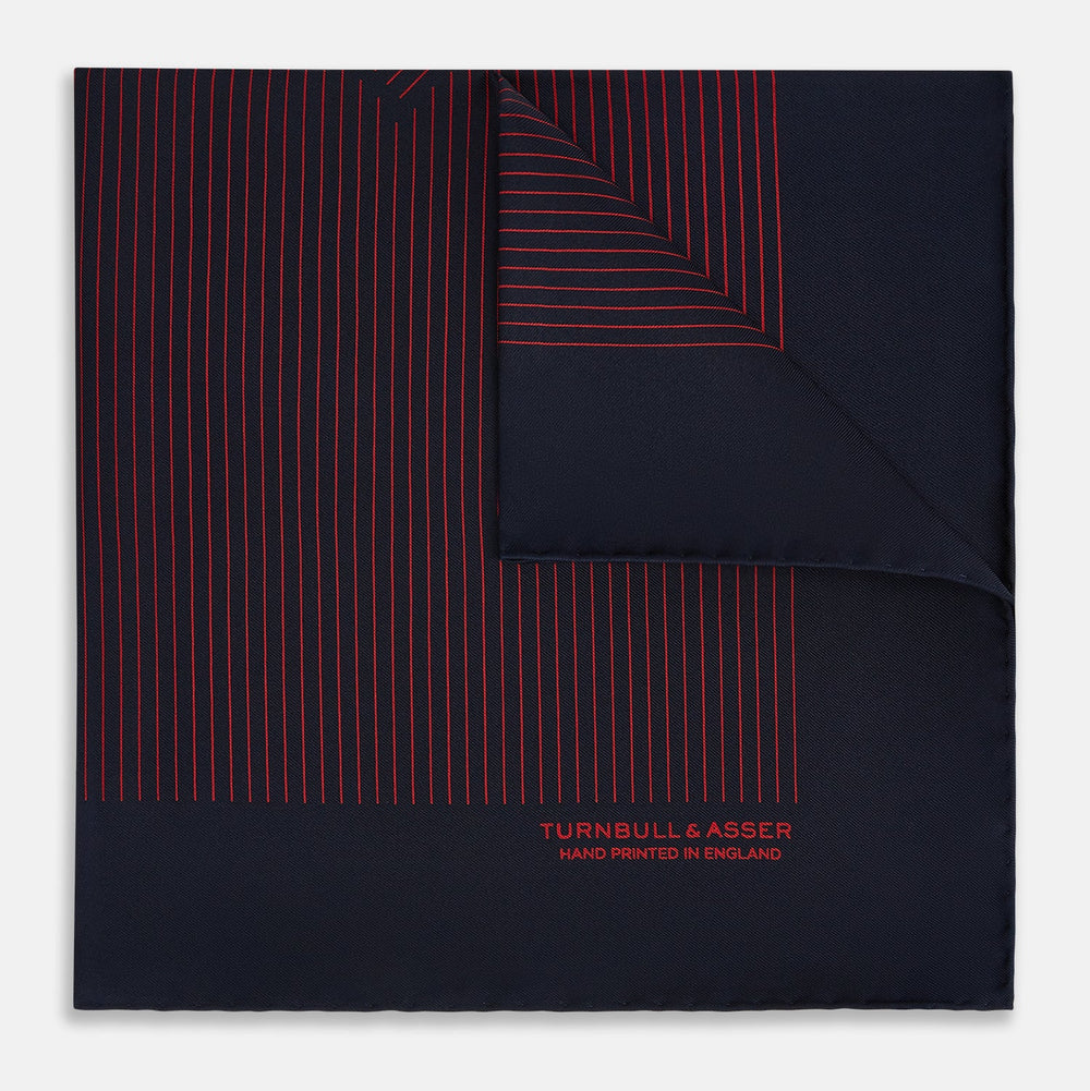 The Turnbull & Asser Navy and Red Stripe Silk Pocket Square features thin red stripes on navy silk, with "HAND PRINTED IN ENGLAND" in red at the corner. A folded corner shows its hand-rolled edges and refined design.