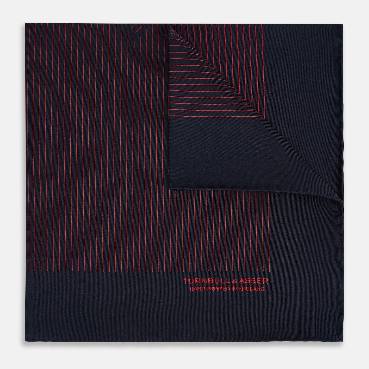 View 1 of product The Turnbull & Asser Navy and Red Stripe Silk Pocket Square features thin red stripes on navy silk, with "HAND PRINTED IN ENGLAND" in red at the corner. A folded corner shows its hand-rolled edges and refined design.