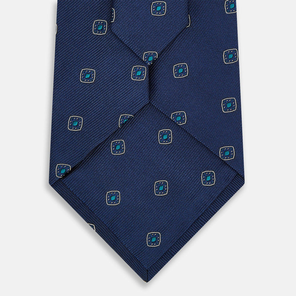 The Turnbull & Asser Navy Geometric Silk Tie showcases a repeating pattern of small light blue and white shapes on a navy base, neatly rolled and displayed against a white background.