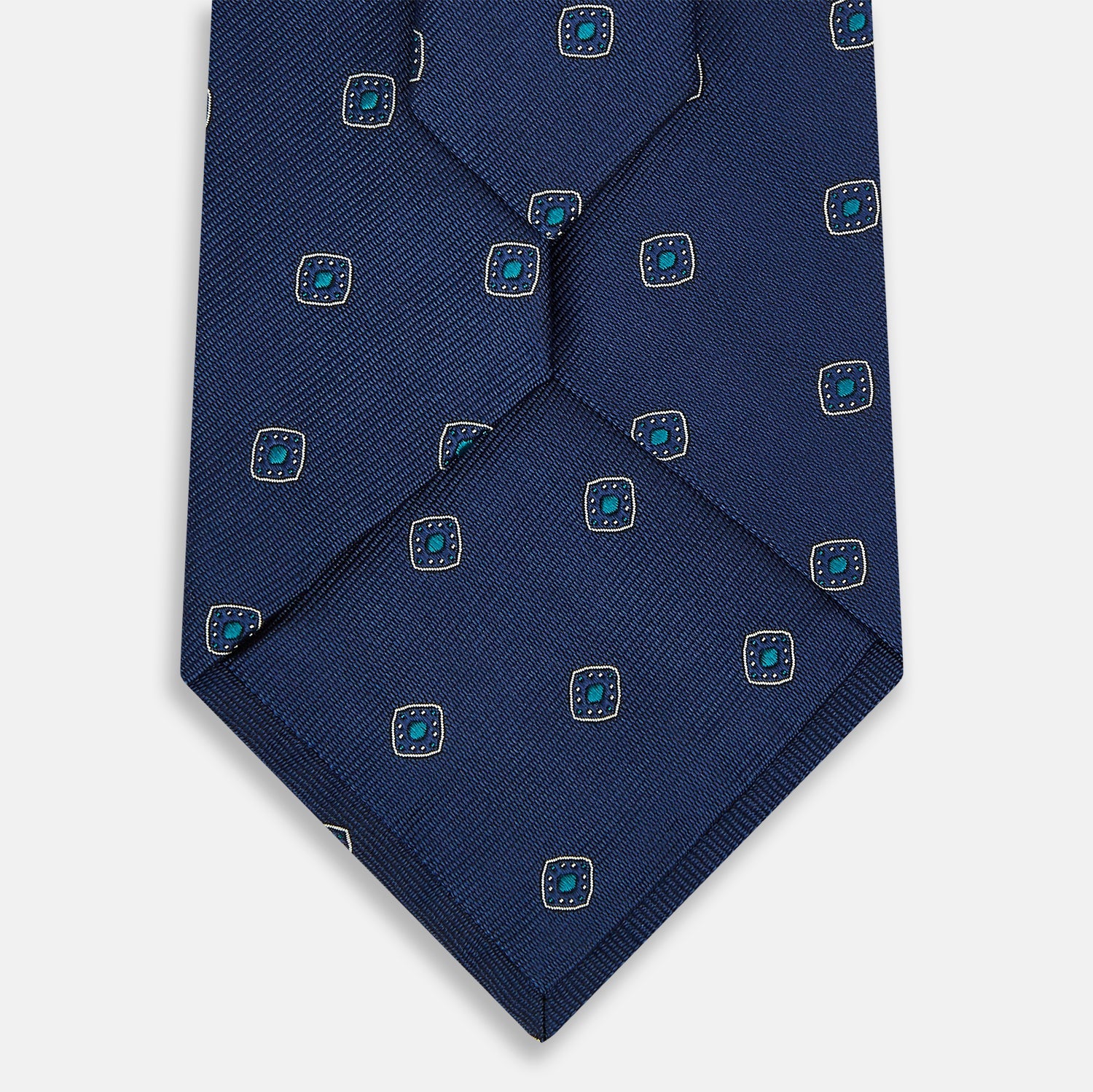 View 3 of product The Turnbull & Asser Navy Geometric Silk Tie showcases a repeating pattern of small light blue and white shapes on a navy base, neatly rolled and displayed against a white background.