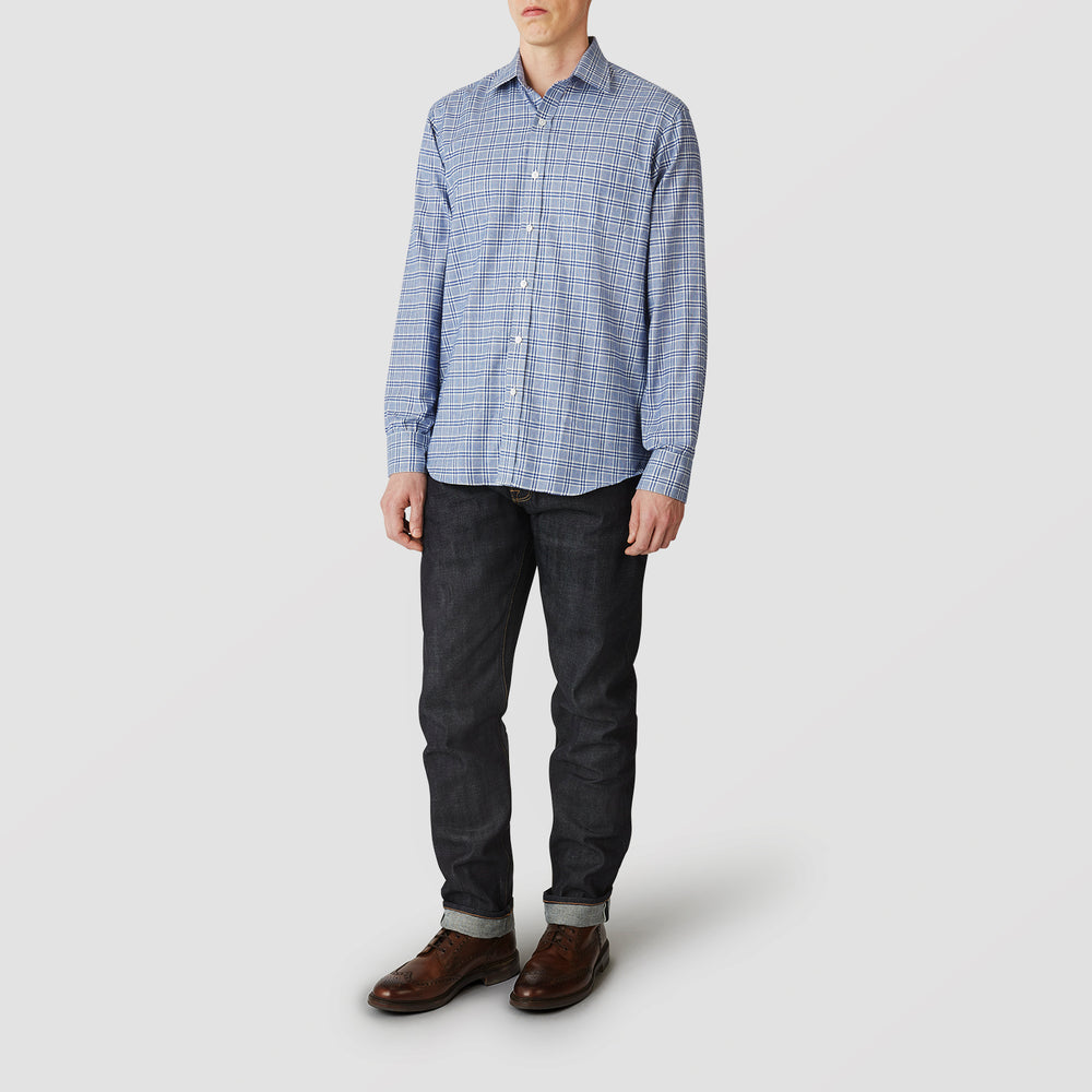 A person in casual attire: a Turnbull & Asser Blue Check Weekend Fit Shirt with Derby Collar and 1-Button Cuffs, dark cuffed jeans, and brown leather shoes, standing against a plain light gray background.