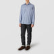 A person in casual attire: a Turnbull & Asser Blue Check Weekend Fit Shirt with Derby Collar and 1-Button Cuffs, dark cuffed jeans, and brown leather shoes, standing against a plain light gray background.