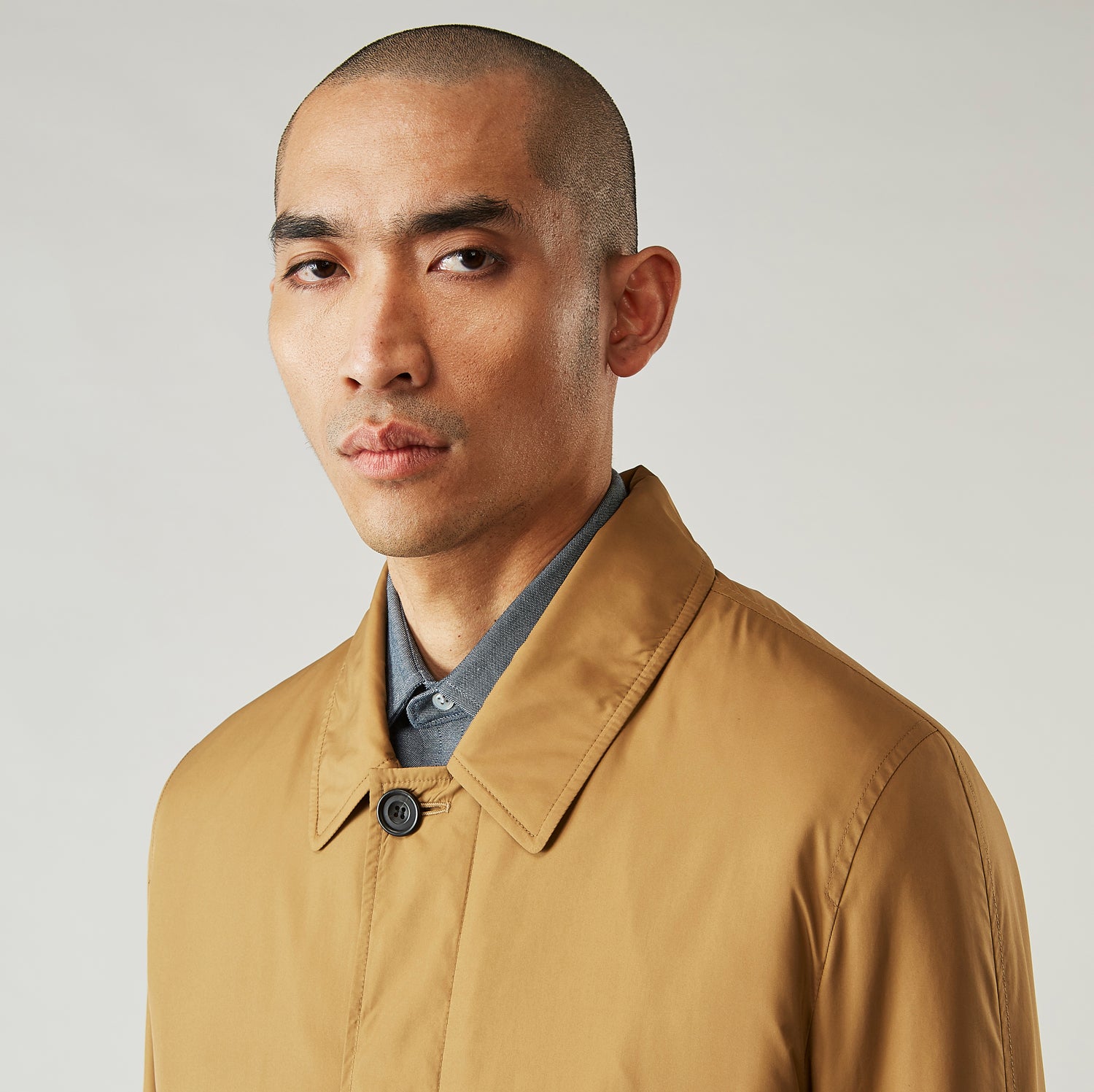 View 5 of product A person wears the Turnbull & Asser Camel Quintern Sealup Raincoat with Detachable Liner, paired with dark pants, standing against a plain light gray background, showing the sleek technical rainwear style with one hand in a pocket.