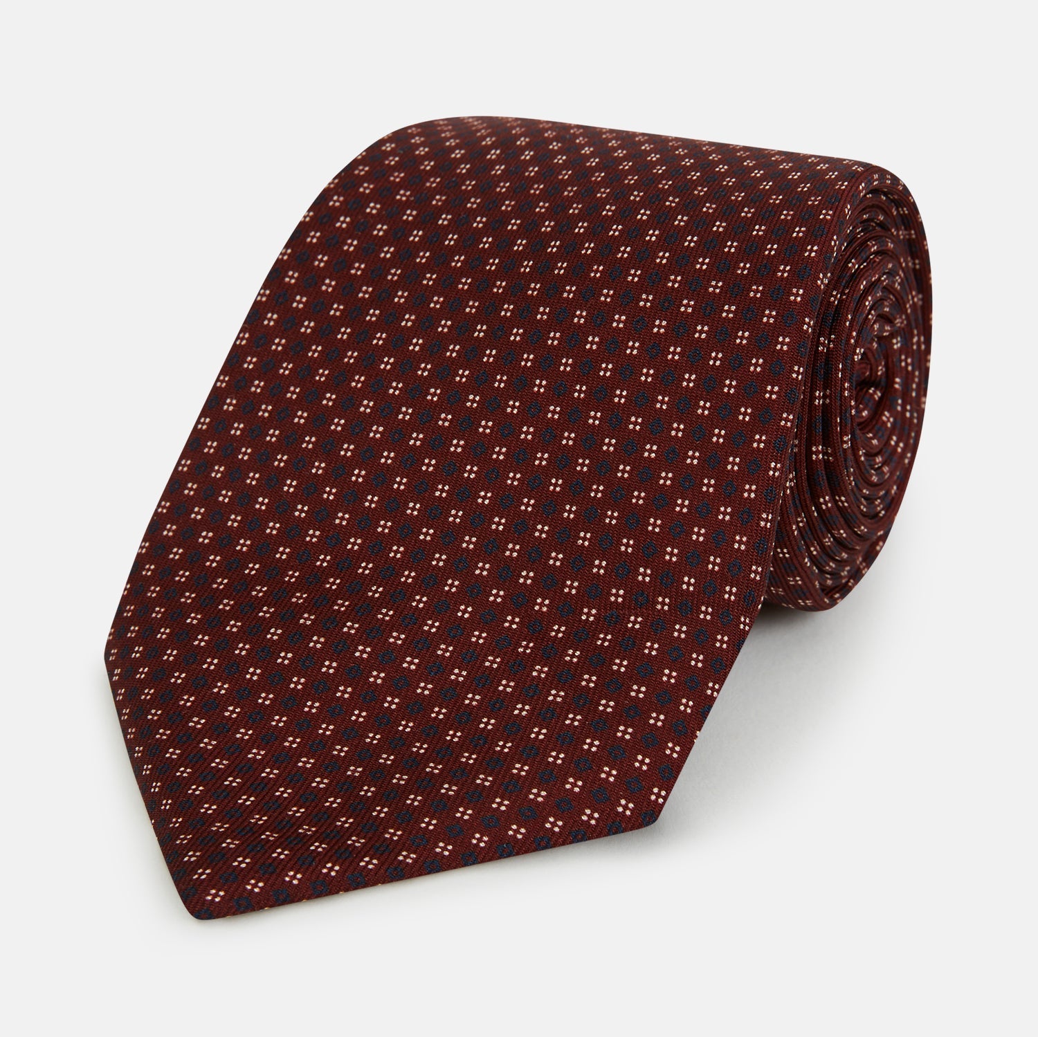 View 1 of product The Burgundy Geometric Motif Silk Tie by Turnbull & Asser features blue and white dots and is displayed neatly rolled on a plain white background.