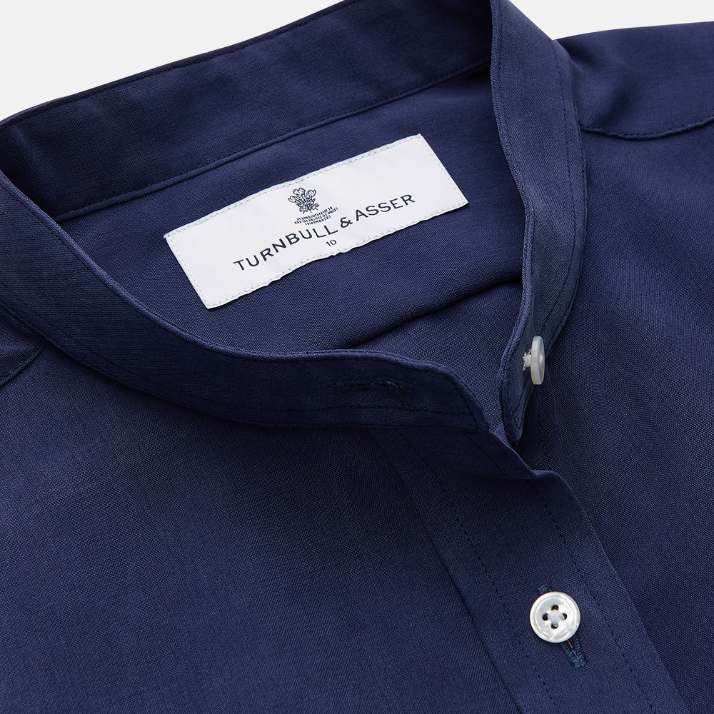 The Turnbull & Asser Blue Soft-touch Silk Lauren Shirt is a navy, long-sleeve button-up with an Igret stand collar, white buttons, curved hem, relaxed fit, and elongated body for a modern look, shown laid flat on a white background.
