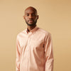 View 6 of product A man faces forward in a relaxed pose against a beige background, wearing the Turnbull & Asser Pink Piece Dyed Weekend Fit Suffolk Shirt with a button-down collar, dark cuffed jeans, and black shoes.