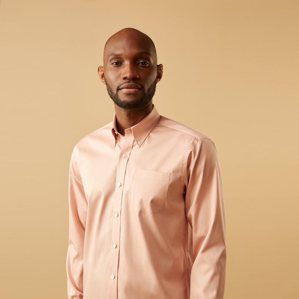 A man faces forward in a relaxed pose against a beige background, wearing the Turnbull & Asser Pink Piece Dyed Weekend Fit Suffolk Shirt with a button-down collar, dark cuffed jeans, and black shoes.