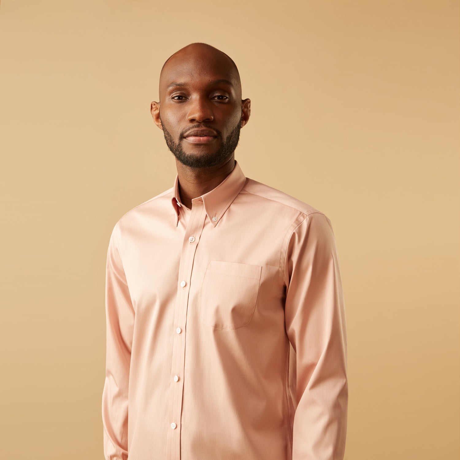 View 6 of product A man faces forward in a relaxed pose against a beige background, wearing the Turnbull & Asser Pink Piece Dyed Weekend Fit Suffolk Shirt with a button-down collar, dark cuffed jeans, and black shoes.