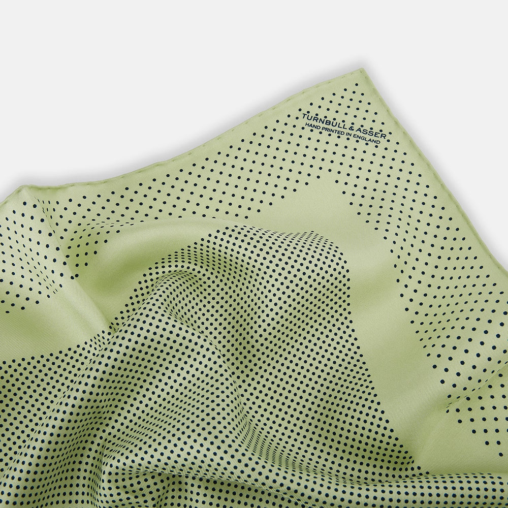 The Melon Green Dot Silk Pocket Square by Turnbull & Asser is handmade in England from silk, featuring small black polka dots. Shown folded with a turned-up corner, it displays both sides of the light green fabric.