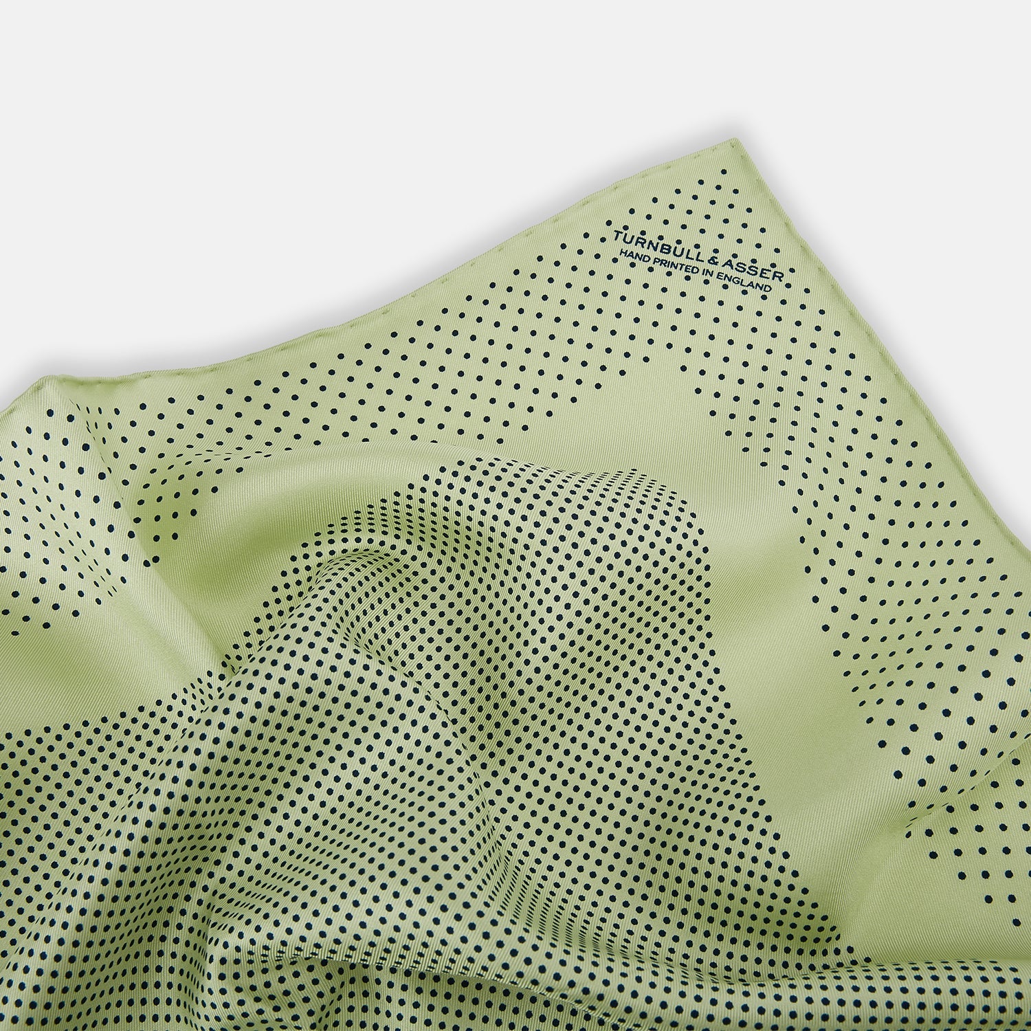 View 2 of product The Melon Green Dot Silk Pocket Square by Turnbull & Asser is handmade in England from silk, featuring small black polka dots. Shown folded with a turned-up corner, it displays both sides of the light green fabric.