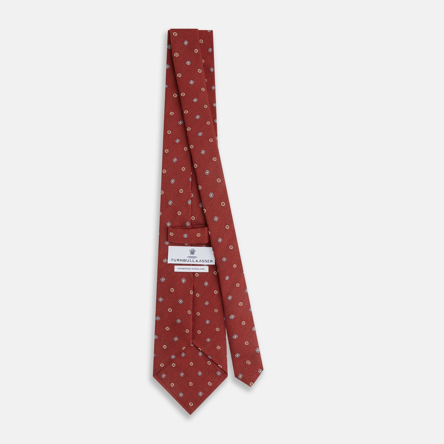 View 4 of product A Turnbull & Asser Red Motif Silk Tie featuring small blue and yellow floral designs with brown geometric shapes is displayed rolled up on a plain white background.