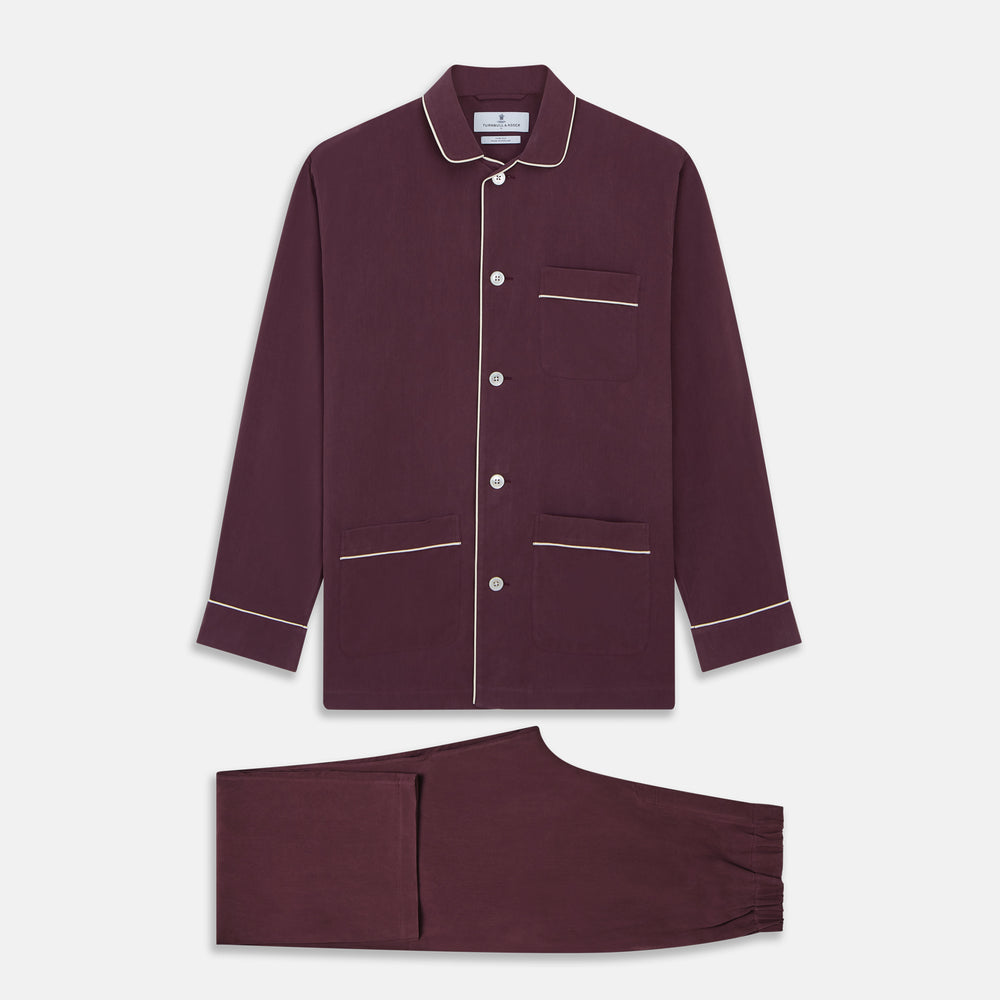 The Turnbull & Asser Plain Plum Silk Modern Pyjama Set, made in England, features white piping, a button-up long-sleeve top with three pockets, and matching pants. The pyjamas are shown against a white background.