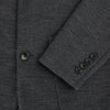 View 4 of product The Turnbull & Asser Graphite Knitted Oxford Blazer for men features a dark gray hue, two buttons, notched lapels, and three front pockets—a modern British-inspired piece styled elegantly against a light background.