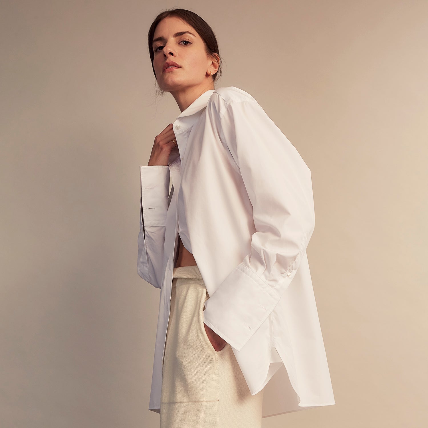View 3 of product A woman with straight brown hair pulled back wears the Turnbull & Asser White Cotton Marlene Shirt, featuring classic menswear details, paired with high-waisted cream trousers against a neutral background.