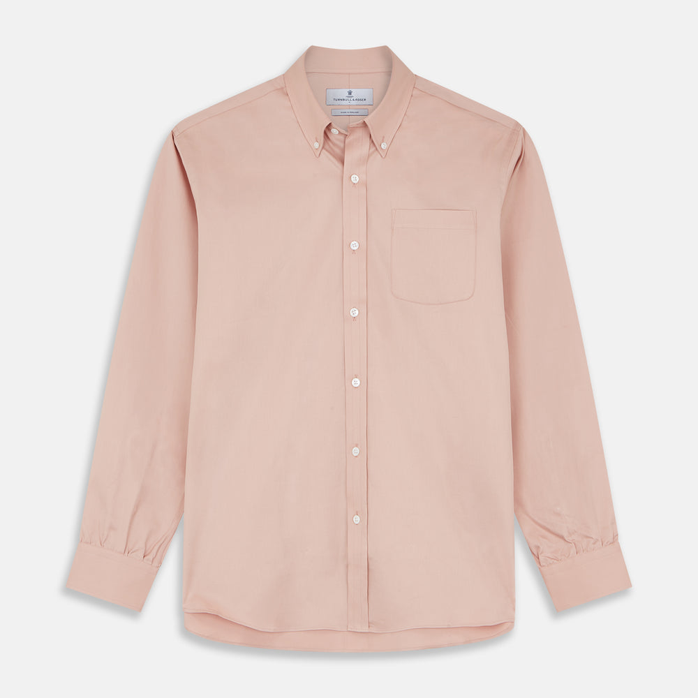 A man faces forward in a relaxed pose against a beige background, wearing the Turnbull & Asser Pink Piece Dyed Weekend Fit Suffolk Shirt with a button-down collar, dark cuffed jeans, and black shoes.