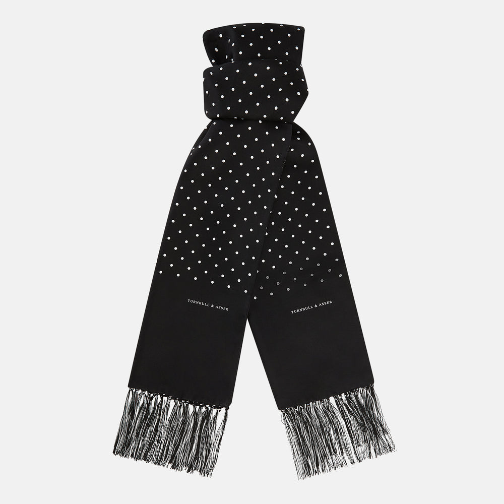 The Turnbull & Asser Black and White Spotted Silk Scarf with Fringes features a classic polka dot design, silver fringe edges, and the brand name printed near each end.