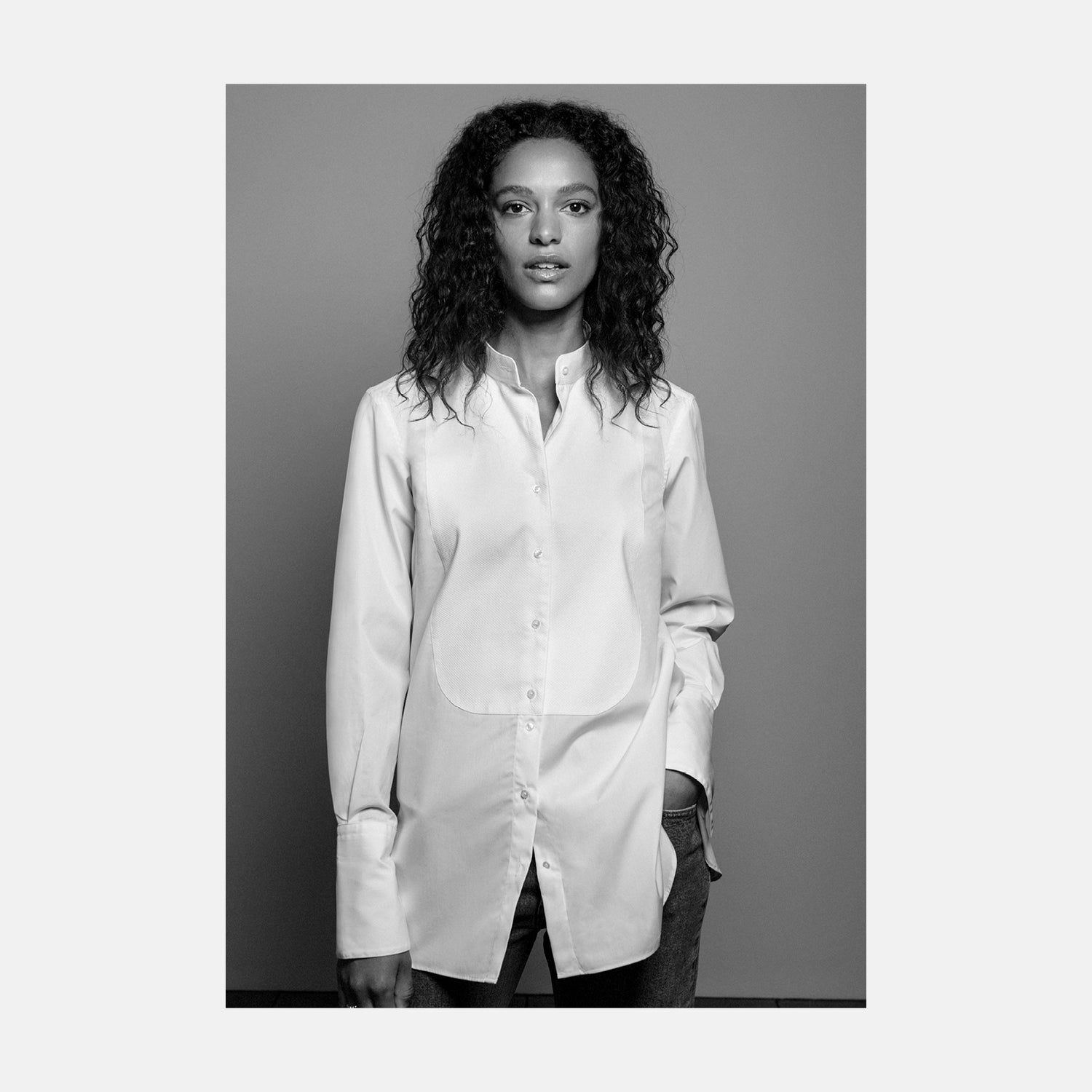 View 1 of product A person with curly hair wears the Turnbull and The Deck White Cotton Minimalist Shirt by Turnbull & Asser, paired with pants. Posed against a plain background, their minimalist silhouette is highlighted in this black and white image.