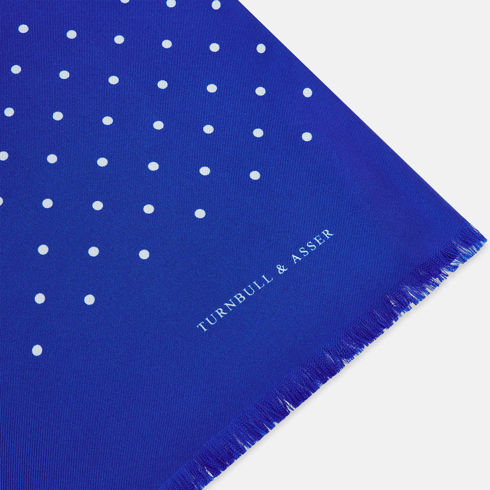 The Turnbull & Asser Royal Blue and White Spotted Silk Scarf features evenly spaced small white dots, fringed edges, and the brand’s name printed near the ends.