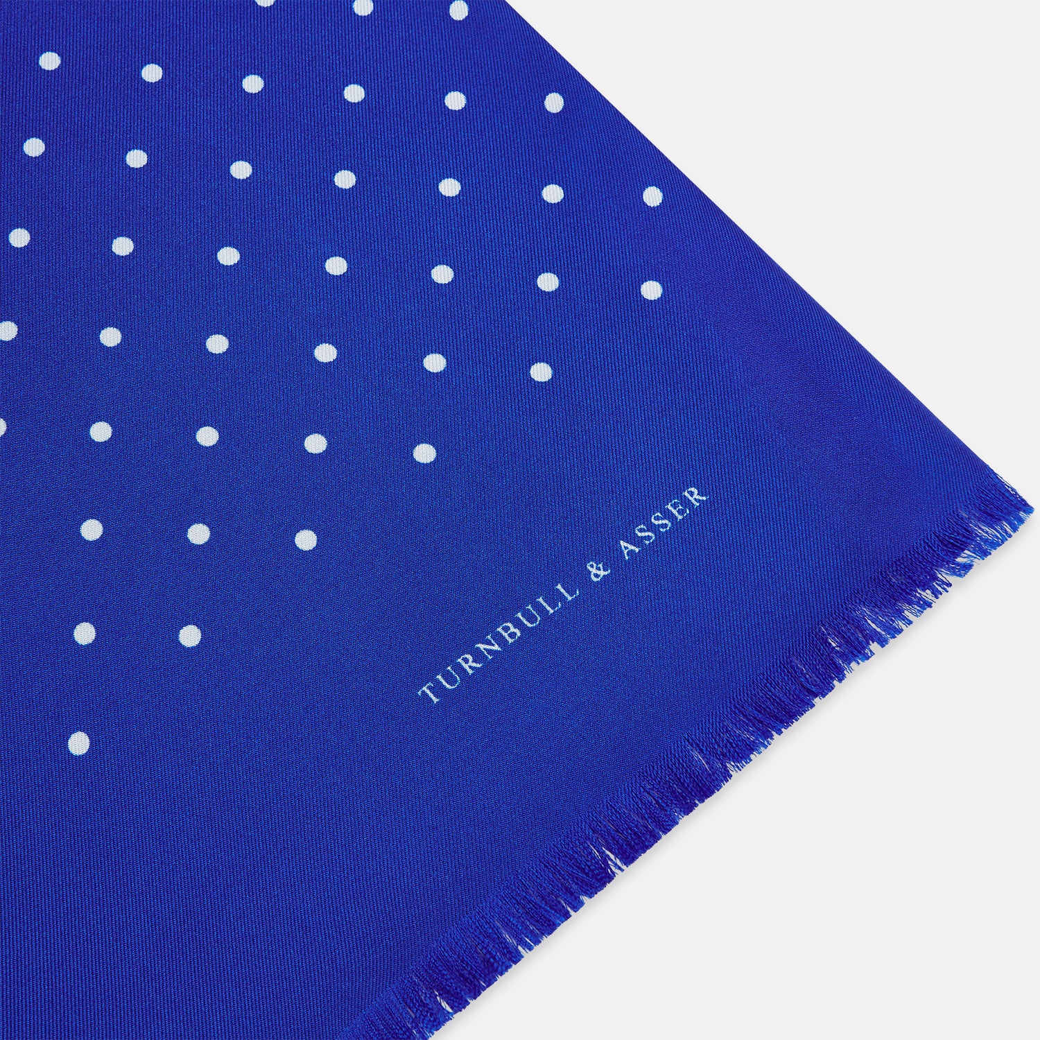View 2 of product The Turnbull & Asser Royal Blue and White Spotted Silk Scarf features evenly spaced small white dots, fringed edges, and the brand’s name printed near the ends.