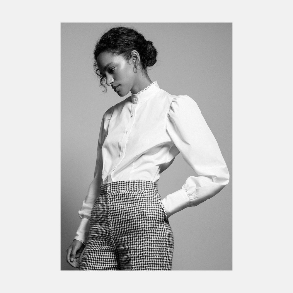 A woman stands in profile, looking down with hands in her pockets, wearing the Turnbull & Asser Turnbull and The Deck White Cotton Expressionist Shirt, paired with high-waisted black-and-white houndstooth pants.