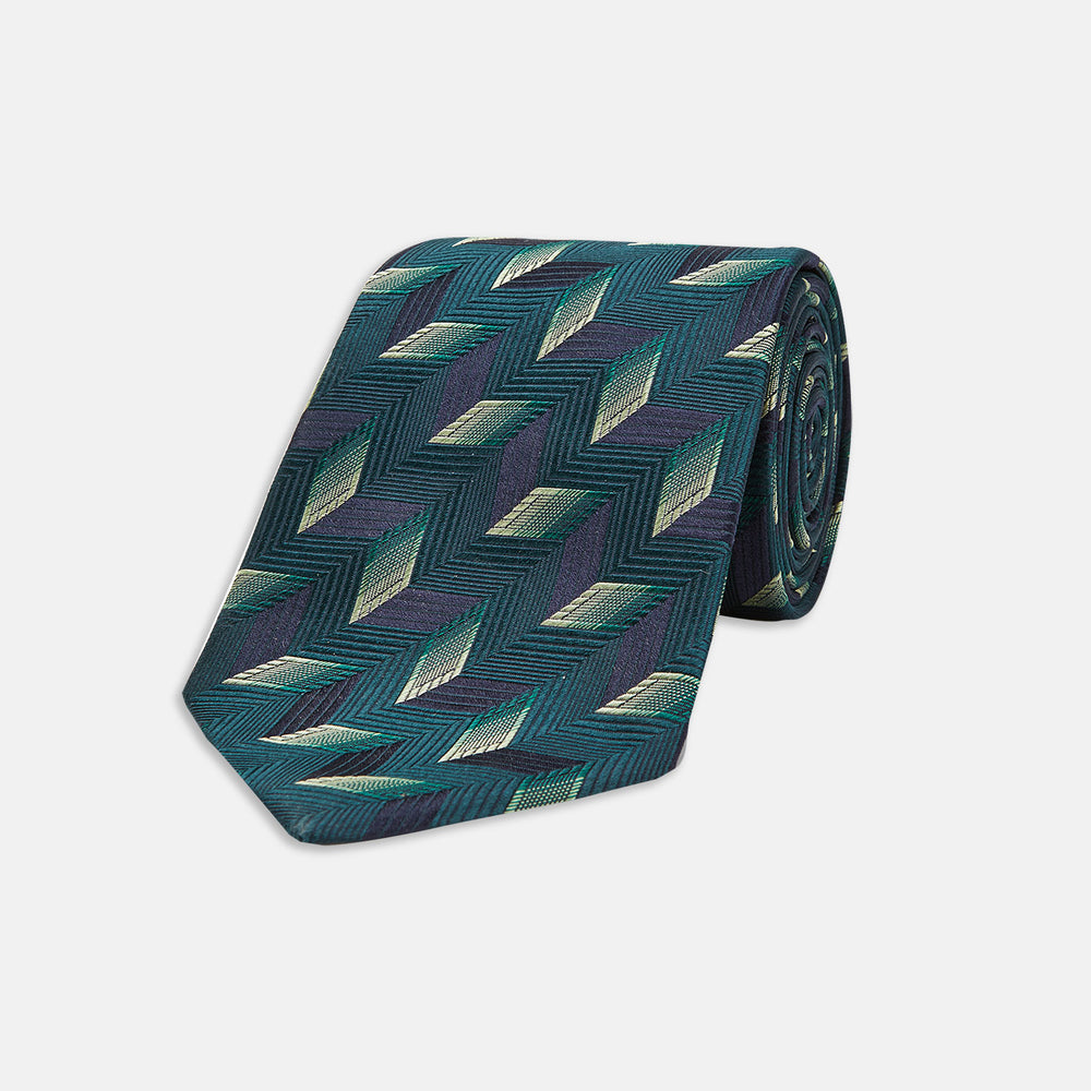 The Turnbull & Asser "The Dark Knight Silk Tie as seen on the Joker" is shown rolled up, featuring a teal zigzag geometric pattern with blue, green, and cream diamonds on a white background.