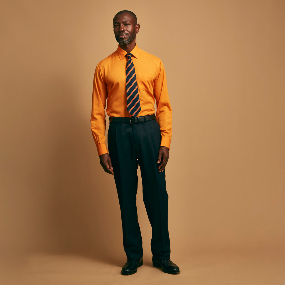 A man stands against a tan background, wearing a Turnbull & Asser Orange Weekend Fit Cotton & Wool Shirt with Derby Collar and 1 Button Cuffs, tucked into dark blue dress pants with a black belt and shoes. He has one hand in his pocket.