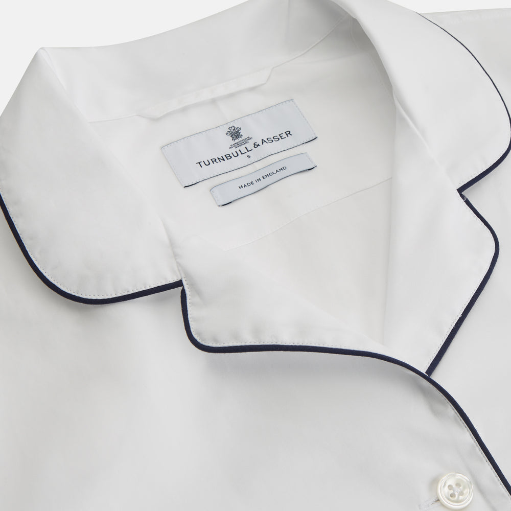 The Turnbull & Asser Women's Harriet Plain White Cotton Pyjama Set features long sleeves, black piping, a chest pocket, and matching pants with elastic waistband and ribbon tie, all displayed on a white background.
