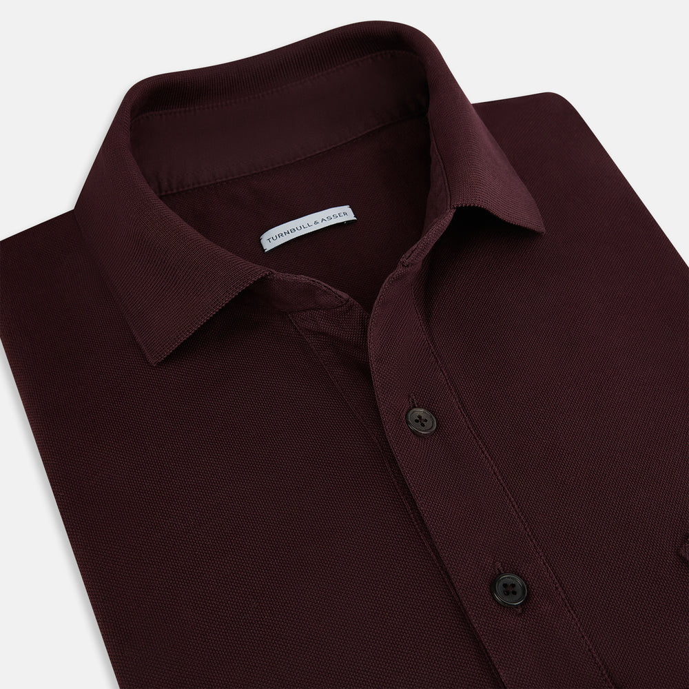 The Turnbull & Asser Burgundy Polo Shirt is crafted from soft cotton jersey with short sleeves, a classic collar, three front buttons, and a small buttoned chest pocket, shown flat on a white background.