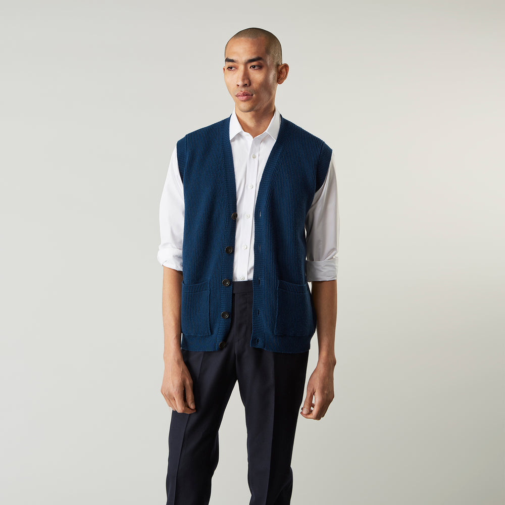 A man with a shaved head wears a Turnbull & Asser Navy Sleeveless Cashmere Cardigan over a white dress shirt with rolled-up sleeves and dark trousers, standing against a plain light gray background.