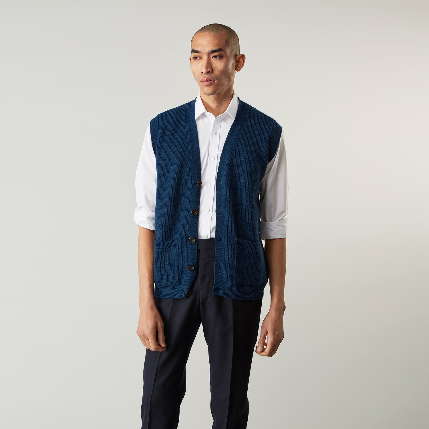 View 3 of product A man with a shaved head wears a Turnbull & Asser Navy Sleeveless Cashmere Cardigan over a white dress shirt with rolled-up sleeves and dark trousers, standing against a plain light gray background.