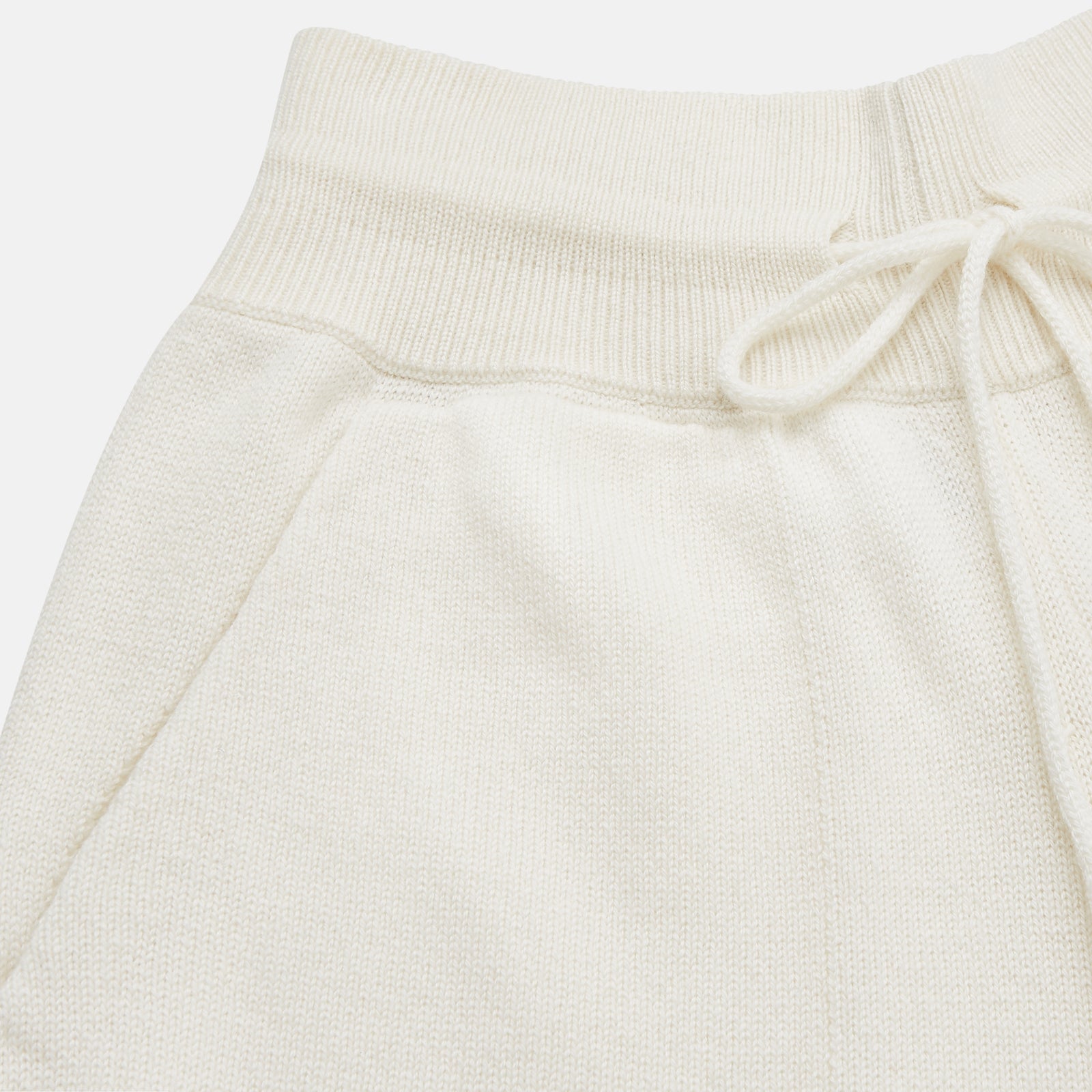 View 5 of product A woman stands barefoot indoors against a neutral wall, wearing Turnbull & Asser Women's Cream Cashmere Knitted Tabitha Bottoms and a light brown sweater. Soft sunlight casts a gentle shadow on the wall beside her.