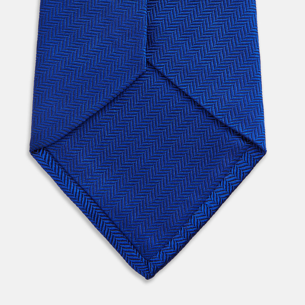 The Turnbull & Asser Plain Herringbone Cruise Tie in bright blue, shown rolled up on a white background, features a subtle chevron pattern—an essential finishing touch for your smart look.