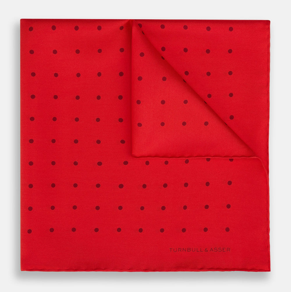 The Turnbull & Asser Red and Burgundy Spot Silk Pocket Square features evenly spaced small spots and the brand logo in the bottom right corner—perfect for formal wear. One corner is folded to show the reverse side.