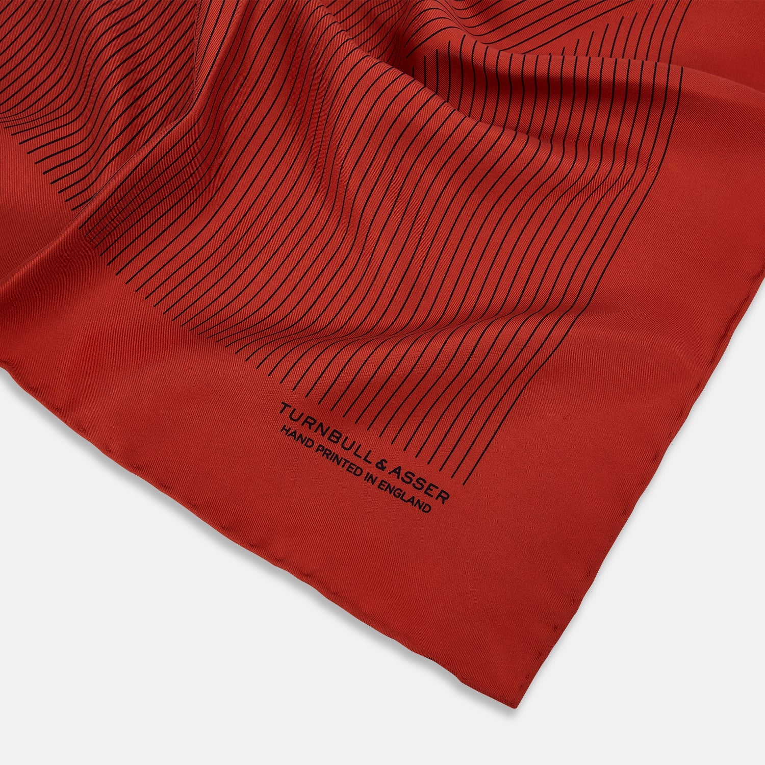 View 2 of product The Red Stripe Silk Pocket Square by Turnbull & Asser features thin black vertical lines and the brand signature in black on one corner, with a neatly folded edge for a touch of classic British style.