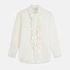 View 2 of product A man in a Cream Silk Regular Fit Ruffle-Front Wilfred Shirt by Turnbull & Asser stands against a dark background, one hand on a chair, gazing thoughtfully to the side.