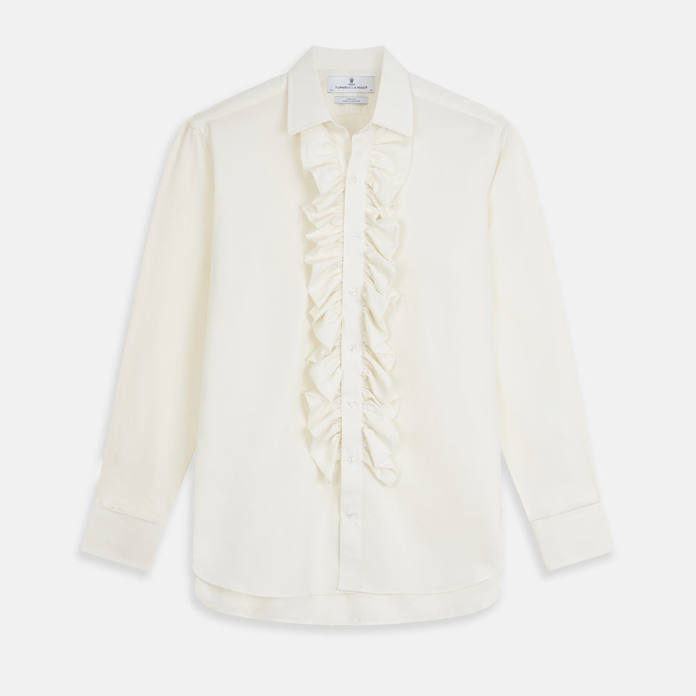 A man in a Cream Silk Regular Fit Ruffle-Front Wilfred Shirt by Turnbull & Asser stands against a dark background, one hand on a chair, gazing thoughtfully to the side.
