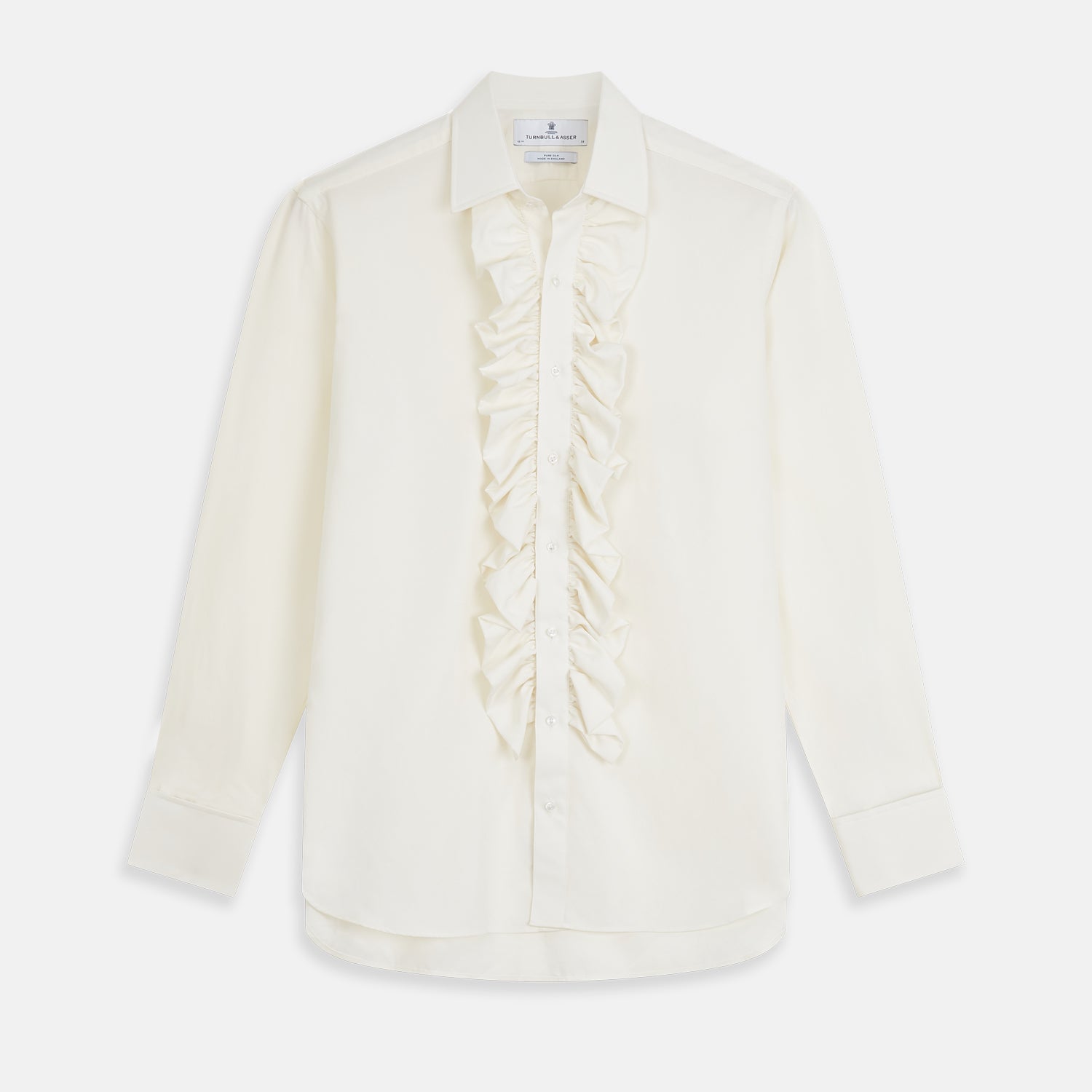 View 2 of product A man in a Cream Silk Regular Fit Ruffle-Front Wilfred Shirt by Turnbull & Asser stands against a dark background, one hand on a chair, gazing thoughtfully to the side.