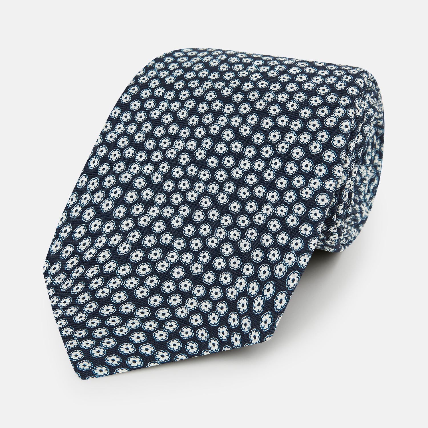 View 1 of product The Turnbull & Asser Navy Flower Patterned Silk Tie is shown rolled up, displaying a repeating floral design with small white and light blue circles on navy, set against a plain white background.