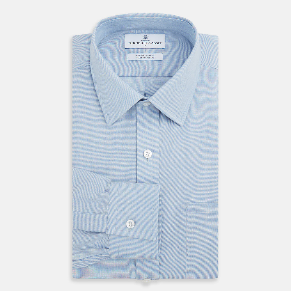 The Turnbull & Asser Pale Blue Cotton Cashmere Derby Collar Richmond Shirt features a soft cotton-cashmere blend, derby collar, white buttons, left chest pocket, and buttoned cuffs, shown laid flat on a white background.