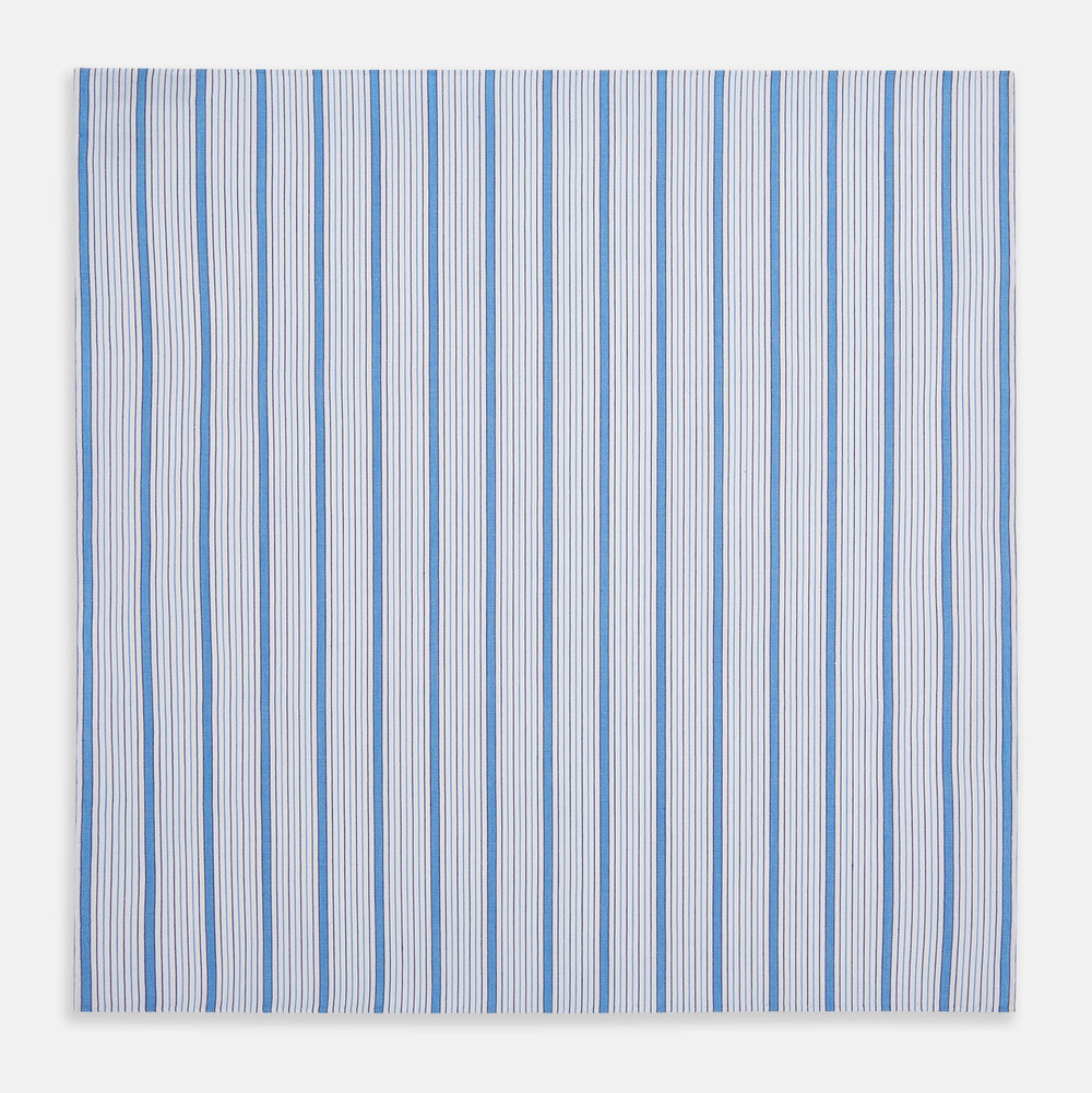 The Hand Rolled Blue Multi Track Stripe Handkerchief by Turnbull & Asser features a square white design with vertical light blue and navy stripes of varying widths—a timeless gentleman’s accessory.