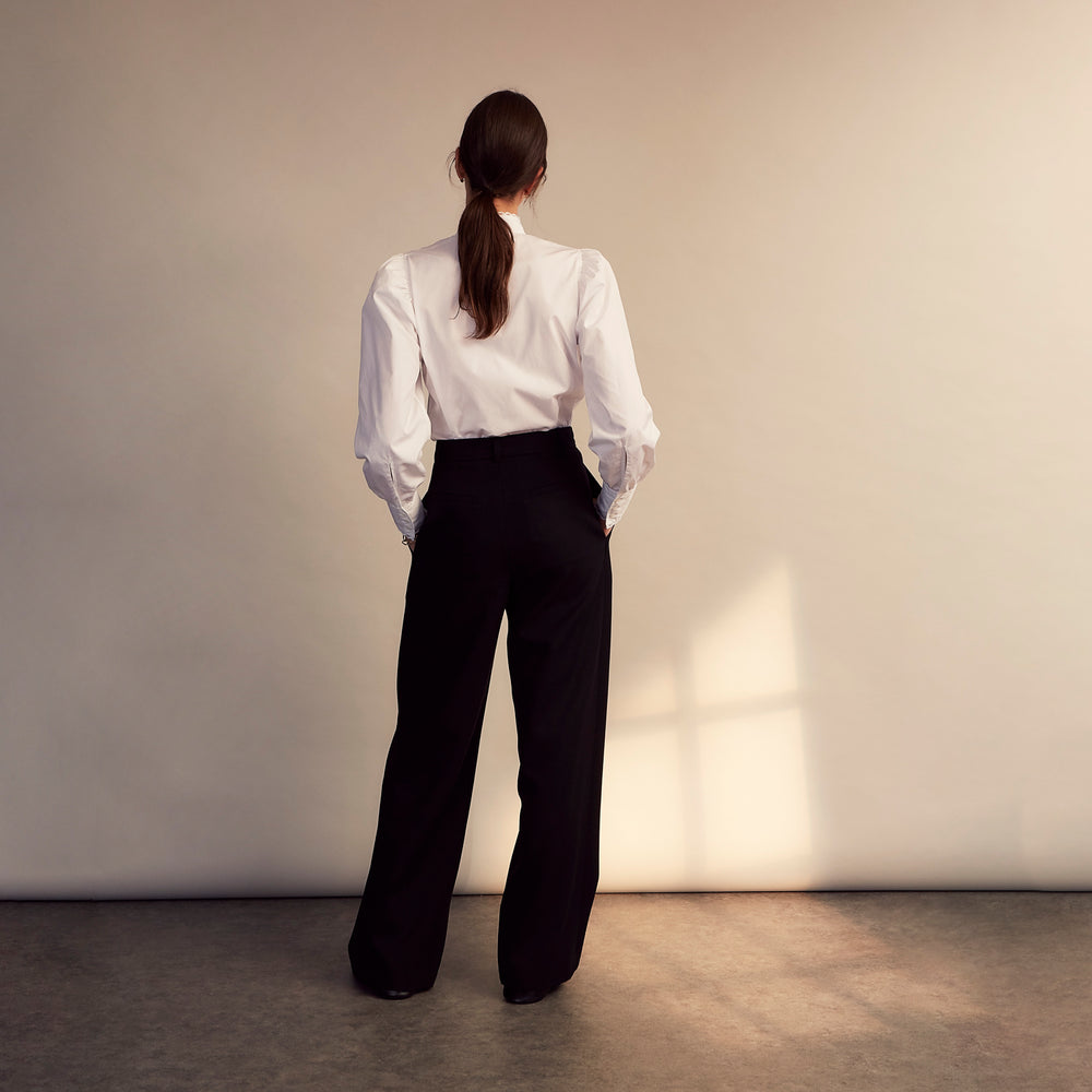 A woman stands in profile, looking down with hands in her pockets, wearing the Turnbull & Asser Turnbull and The Deck White Cotton Expressionist Shirt, paired with high-waisted black-and-white houndstooth pants.